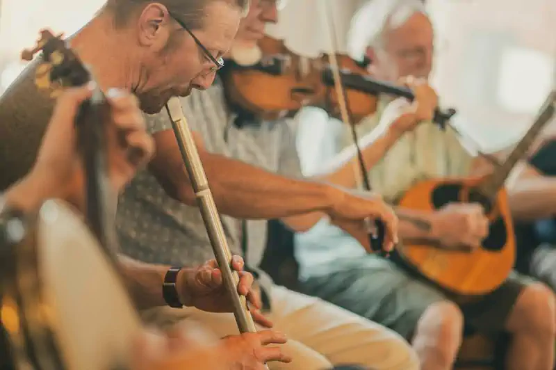 A group of musicians play traditional instruments, including a flute and violins, in a close setting. The scene conveys warmth and camaraderie.