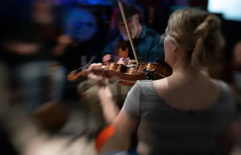 A woman with blurred surroundings plays the violin passionately in a dimly lit room, creating an intimate and immersive musical atmosphere.