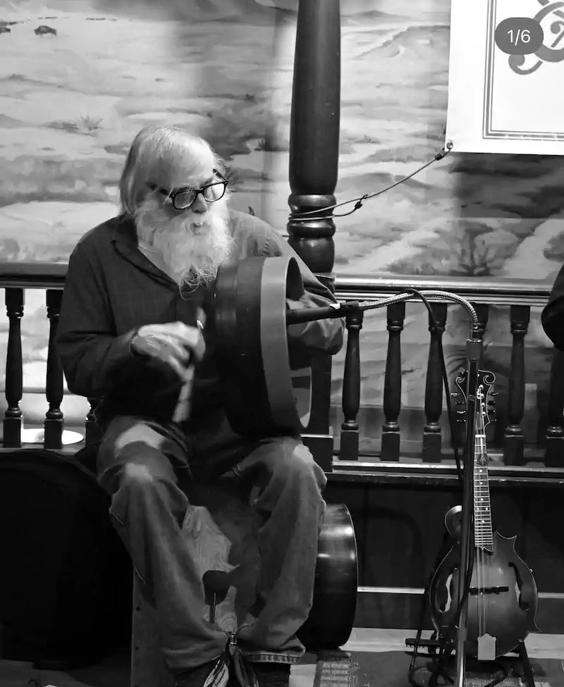 An elderly man with a long white beard and glasses plays a drum with focus and passion. A mandolin is propped nearby. Warm, nostalgic ambiance.