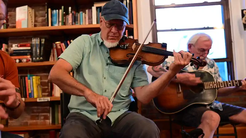 A man in a blue cap plays a violin passionately, while another man with white hair strums a guitar in the background. They are in a cozy room with bookshelves, creating an intimate, musical atmosphere.