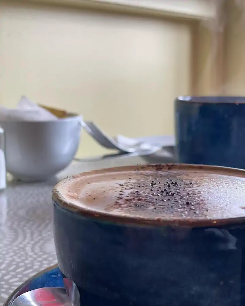 A close-up of a steaming cappuccino in a navy cup, topped with cocoa, on a patterned surface. A sugar bowl and spoon are blurred in the background. Cozy ambiance.