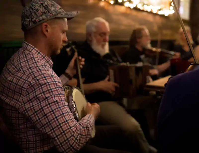 A cozy pub setting features musicians playing various instruments. In the foreground, a man in a plaid shirt plays the banjo. Warm lighting and a relaxed, joyful atmosphere.