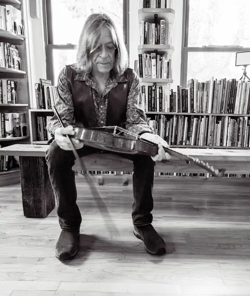 Black and white image of a person with long hair and glasses sitting on a bench, holding a violin. Surrounded by bookshelves, they appear thoughtful.