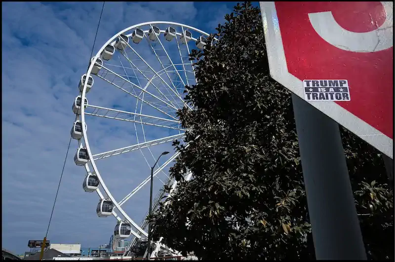 Ferris wheel against a cloudy blue sky, partially obscured by leafy trees. A red sign in the foreground has a sticker reading “TRUMP IS A TRAITOR.”
