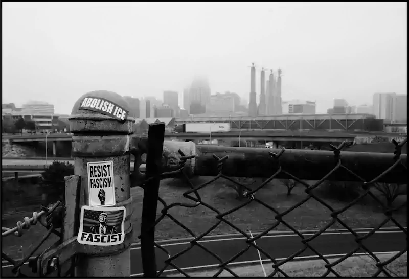 Balck and white cityscape with a foggy skyline in the background. Foreground shows a fence post with stickers reading "Abolish ICE," "Resist Fascism," and "Fascist."