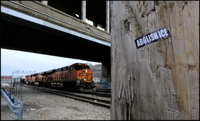 A freight train moves under a bridge on a dreary day. In the foreground, a weathered pole displays a sticker reading "ABOLISH ICE," conveying a political message.