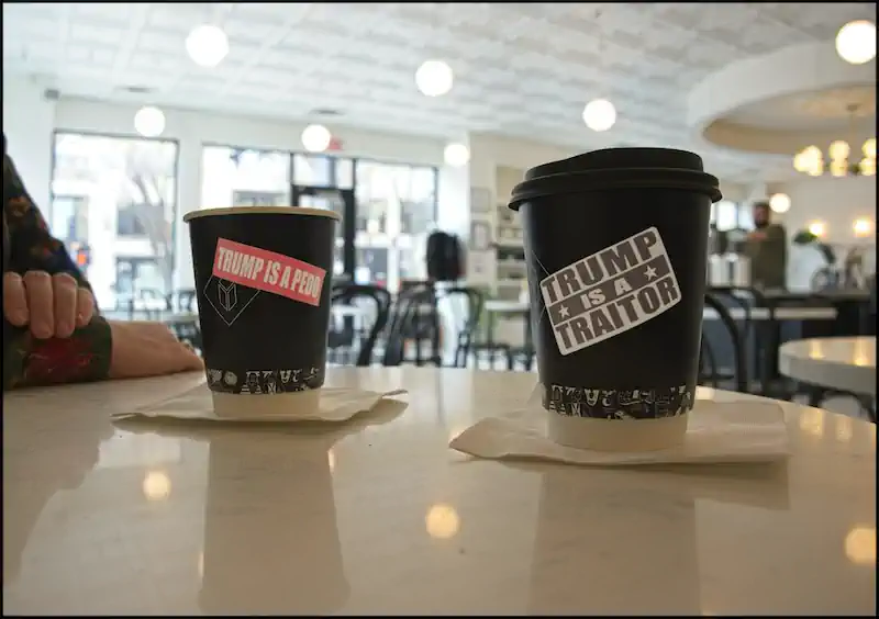 Two coffee cups with controversial political stickers in a bright cafe. One says "Trump is a Pedo," and the other, "Trump is a Traitor." Mood is tense.