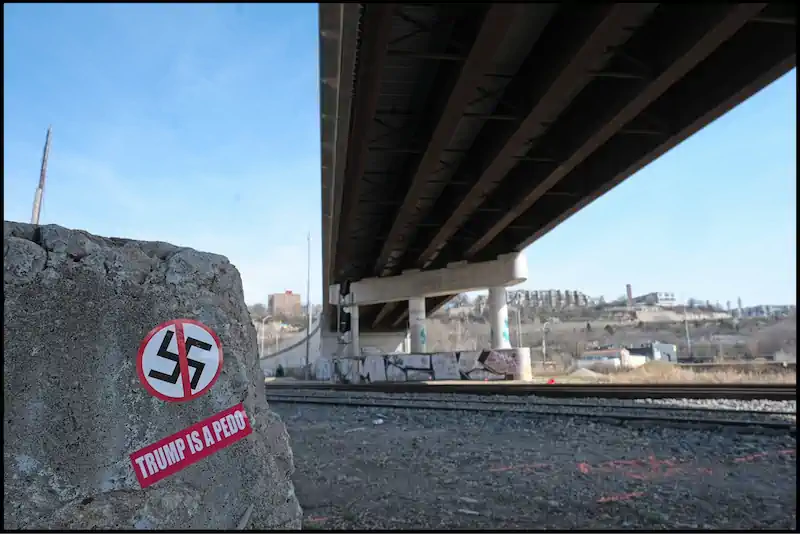 Graffiti under a bridge shows an anti-Trump message and a crossed-out swastika. The scene is urban and desolate, conveying rebellion and dissent.