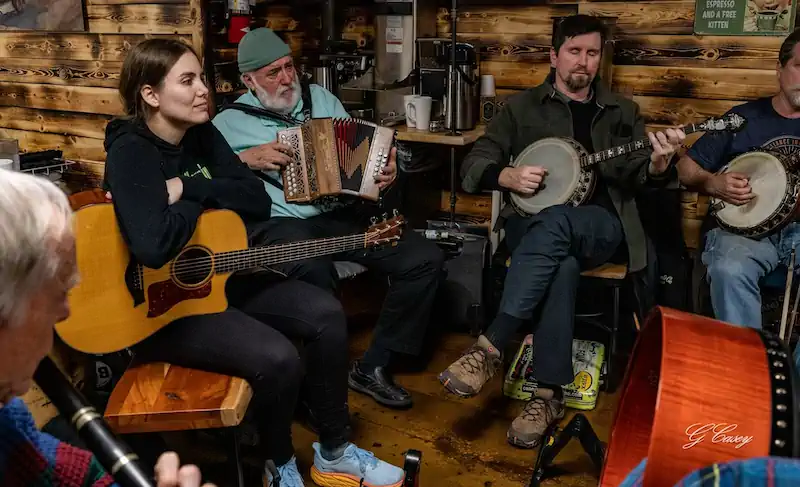 This photo of Turlach Boylan playing the Banjo depicts a cozy indoor gathering with musicians playing various instruments, including a guitar, accordion, and banjo, creating a warm and lively atmosphere.