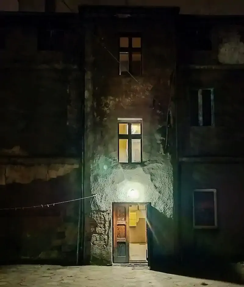 Dimly lit, old building facade at night with a single glowing entrance, casting shadows on a worn cobblestone courtyard, evoking a mysterious atmosphere.