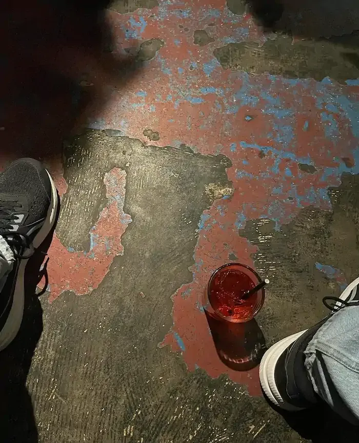 Worn sneakers next to a red drink on a textured, distressed floor with blue and red paint. The scene feels casual and laid-back.