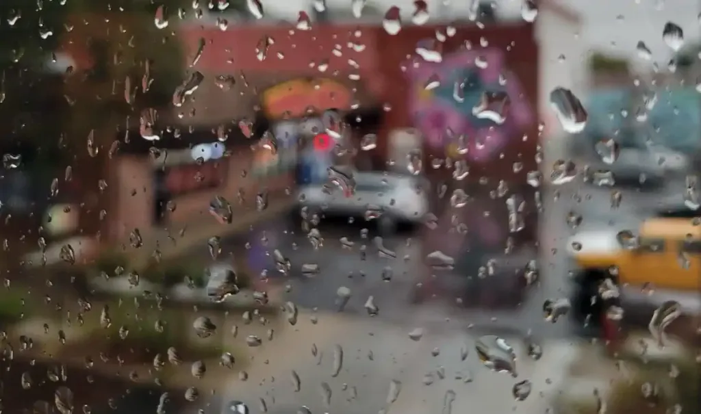 Raindrops on a window blur a street scene, creating a soft and dreamy effect. The blurred background shows red buildings and a yellow car.
