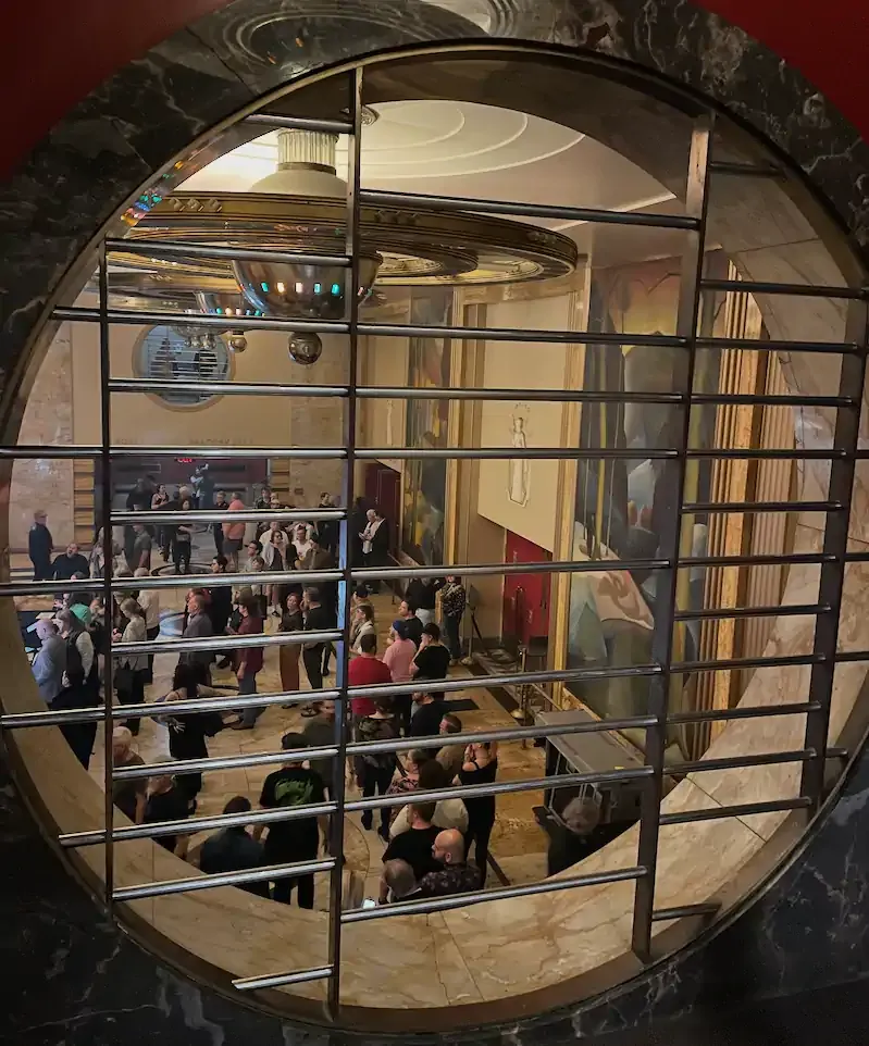 View through circular window bars showing a bustling art deco lobby with elegant murals. People are mingling, creating a lively, upscale atmosphere.
